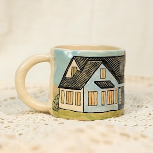 Könnte beinhalten: Eine Keramiktasse mit handgemaltem Hausdesign. Die Tasse zeigt ein hellblaues Haus mit schwarzem Dach und gelben Fenstern. Der Griff und der Rand der Tasse sind cremefarben. Die Tasse steht auf einer weißen Spitzentischdecke.