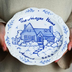 May include: A white ceramic plate with a blue and white design of a house, trees, and a fence. The text "The Magee Home August 2023" is written on the plate.