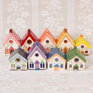 Puede incluir: Una colección de casas en miniatura de cerámica coloridas. Cada casa presenta una combinación de colores única, incluyendo rosa, rojo, naranja, amarillo, morado, azul y verde. Las casas tienen detalles arquitectónicos y acentos decorativos.