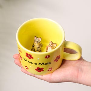 K&ouml;nnte beinhalten: Eine gelbe Keramiktasse mit Henkel, verziert mit roten Blumen und wei&szlig;en Punkten. Im Inneren der Tasse befinden sich zwei kleine Katzenfiguren. Auf der Tasse steht der Text "Gina e Mela".