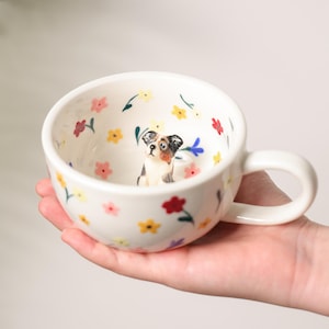 May include: A white ceramic mug with a handle, featuring a floral pattern and a small dog figurine inside. The flowers are red, yellow, pink, and blue. The mug is held in a hand and has a glossy finish. The mug is suitable for tea or coffee.