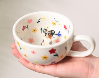 May include: White ceramic mug with a handle, decorated with colorful flowers and a small dog figurine inside. The mug has a smooth, glossy finish and is held in a hand. The flowers are red, yellow, pink, and blue.