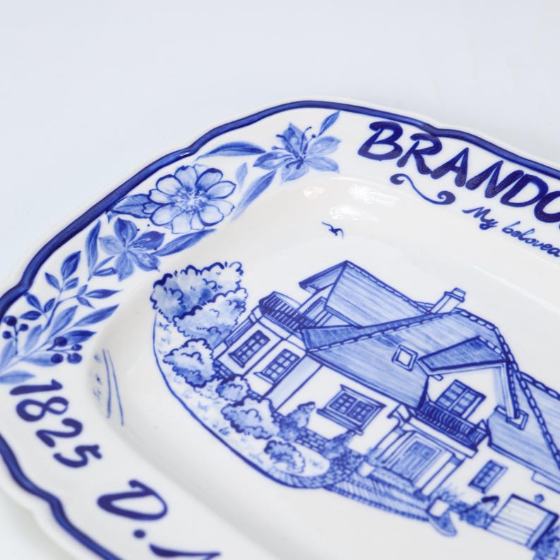 May include: A white ceramic serving dish with a blue floral border and a detailed blue illustration of a house. The dish has the text "BRANDON" and "1825 D." in blue.