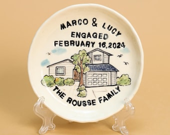 Op de afbeelding: Een wit keramiek bord met een handgeschilderd ontwerp van een huis, bomen en vogels. Het bord is beschreven met de tekst "Marco & Lucy Verloofd Februari 16, 2024 De Familie Rousse".