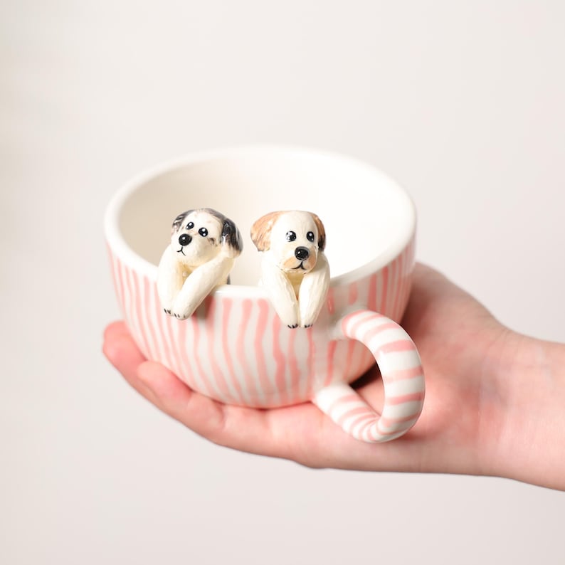 K&ouml;nnte beinhalten: Eine Keramiktasse mit rosa-wei&szlig; gestreiftem &Auml;u&szlig;eren, mit zwei kleinen Hundefiguren, die auf dem Rand sitzen. Die Tasse hat einen passenden gestreiften Griff und eine wei&szlig;e Innenseite. Die Hunde haben schwarze und braune Markierungen.