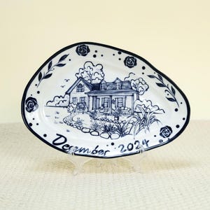 May include: White ceramic platter with a blue and white illustration of a house and garden scene. The platter has a dark blue border with floral and leaf accents. The words "December 2024" are written on the platter.