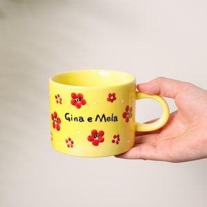 K&ouml;nnte beinhalten: Eine gelbe Keramiktasse mit roten Blumenmustern und dem Text "Gina e Mela". Die Tasse, die von einer Hand gehalten wird, hat einen abgerundeten Henkel. Die Blumen sind rot mit wei&szlig;en Akzenten.