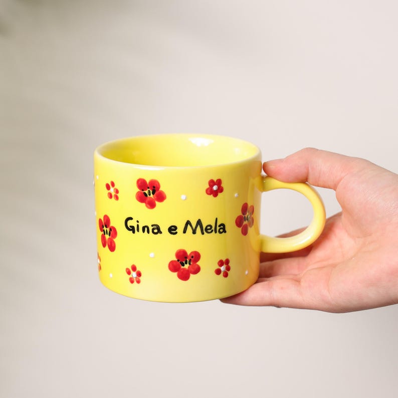 K&ouml;nnte beinhalten: Eine gelbe Keramiktasse mit roten Blumenmustern und dem Text "Gina e Mela". Die Tasse, die von einer Hand gehalten wird, hat einen abgerundeten Henkel. Die Blumen sind rot mit wei&szlig;en Akzenten.