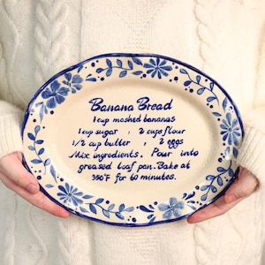 Peut inclure: Plat ovale en céramique crème avec une bordure florale bleue. L'assiette présente une recette de pain aux bananes manuscrite. La recette comprend les ingrédients et les instructions de cuisson.