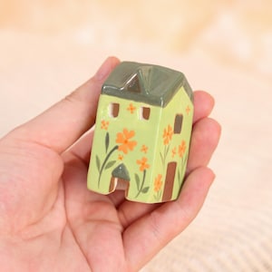 May include: A small, ceramic house figurine held in a hand. The house is painted light green with a dark green roof and brown door and window frames. Orange flowers and green leaves decorate the walls. The figurine has a whimsical, handcrafted appearance.