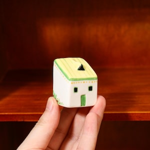 Puede incluir: Una pequeña figura de casa de cerámica blanca con techo amarillo y ribete verde. La casa tiene una puerta verde, ventanas cuadradas negras y una chimenea triangular negra. La figura se sostiene en una mano.