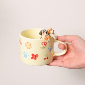 K&ouml;nnte beinhalten: Eine hellgelbe Keramiktasse mit Henkel, verziert mit bemalten Blumen, Pfotenabdr&uuml;cken und Essensillustrationen. Zwei kleine Hundefiguren sitzen auf dem Rand. Die Tasse wird von einer Hand gehalten.