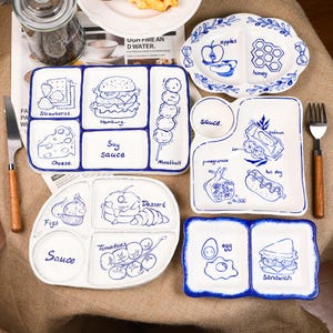 Könnte beinhalten: Eine Sammlung weißer Keramikplatten mit blau handgezeichneten Illustrationen von Lebensmitteln. Die Platten umfassen Abschnitte für Erdbeeren, Käse, Hamburger, Fleischbällchen, Äpfel, Honig, Feigen, Tomaten, Eier und Sandwiches. Einige Platten haben das Wort "Sauce" darauf geschrieben.