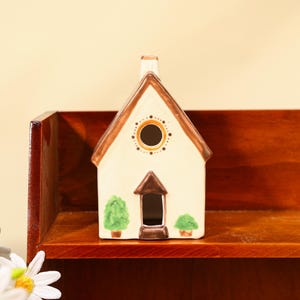 Könnte beinhalten: Eine cremefarbene Keramikhausfigur mit braunem Dach und Schornstein. Das Haus hat ein kreisförmiges Fenster mit gepunkteten Akzenten und eine kleine Tür. Zwei grüne, getopfte Bäume flankieren die Tür. Die Figur steht auf einem Holzregal.
