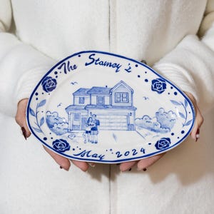 Puede incluir: Un plato de servir ovalado de cerámica blanca con un diseño pintado a mano en azul y blanco. El diseño presenta una casa, dos figuras y el texto "The Stanleys' May 2024".