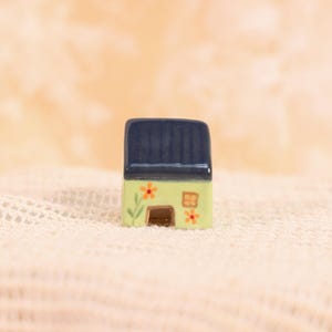 May include: A miniature ceramic house with a dark blue roof and light green walls. The walls are decorated with orange flowers and a small brown door. The house is set against a soft, textured background.