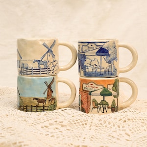 May include: Four ceramic mugs stacked together, each with a unique hand-painted design. The mugs feature scenes of windmills, horses, and quaint shops with text that reads "Bread" and "Cheese". The mugs have a cream-coloured base and curved handles.