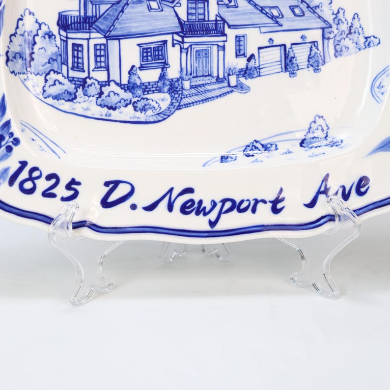 May include: White rectangular ceramic plate with a blue illustration of a house and the text "1825 D. Newport Ave." The plate has a scalloped edge and is supported by a clear plastic stand.