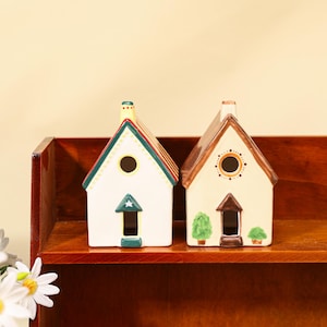 Puede incluir: Dos figuras de casa de cerámica. Una es blanca con un tejado verde y rojo, un diseño de estrella y flecha y una ventana redonda. La otra es beige con un tejado marrón, una ventana redonda y una puerta con árboles verdes. En una estantería de madera.