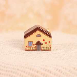 May include: A miniature ceramic house with a brown roof and yellow walls. The house features a blue window, a brown door, and red flower accents. The house is decorated with a scalloped trim and a small chimney.