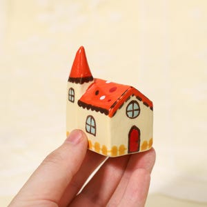 May include: A small ceramic house figurine featuring a cream-colored base, a bright red roof with white polka dots, a pointed tower, a red front door, and painted blue-framed windows. The structure includes a yellow fence detail at the bottom.