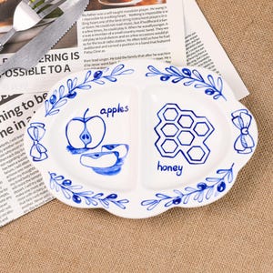 May include: White ceramic serving dish with a blue floral design and two compartments. One side features an apple illustration with the word "apples," and the other a honeycomb with the word "honey."