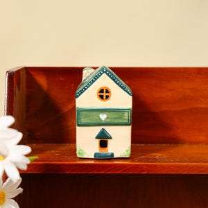 Op de afbeelding: Een keramieken huisfiguur met een crèmekleurig lichaam, groen dak en accenten. Het huis heeft een rond raam met een kruis, een groene lade met een wit hart en een kleine deur. Geplaatst op een houten plank.
