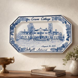 May include: An octagonal white ceramic serving platter featuring a blue hand-painted illustration of Crane Cottage on Jekyll Island. The border is decorated with blue floral patterns, and the text reads Crane Cottage and August 14, 2021.