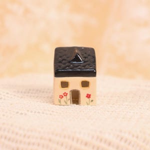 May include: A miniature ceramic house with a black roof and beige walls. The house has two windows and a door, with red flower accents. The house is a decorative item.