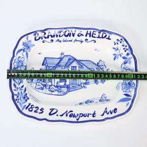 May include: A white ceramic serving platter with a blue floral border and a hand-painted house scene. The platter is approximately 12 inches long. Text includes "BRANDON & HEIDI My beloved family" and "1825 D. Newport Ave."