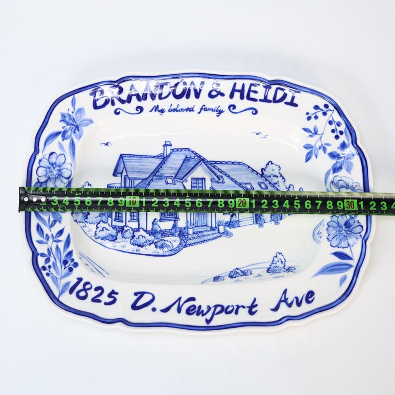 May include: A white ceramic serving platter with a blue floral border and a hand-painted house scene. The platter is approximately 12 inches long. Text includes "BRANDON & HEIDI My beloved family" and "1825 D. Newport Ave."