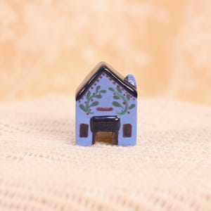 May include: A miniature blue ceramic house with a black roof and door. The house is decorated with green and red floral accents. The small house has a chimney and two small windows. The house is sitting on a white surface.