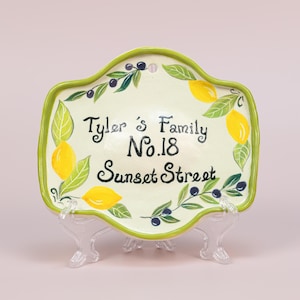 以下が含まれることがあります： レモン、オリーブ、そして「Tyler's Family No.18 Sunset Street」の文字が描かれた、薄緑色の背景のセラミック製の家の看板。看板は緑色の縁取りがあり、透明なプラスチック製のスタンドに飾られています。