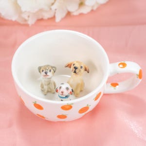 May include: A white ceramic mug with a handle, decorated with orange fruit designs. Inside the mug are three small ceramic dog figurines: a dalmatian, a brown and white dog, and a yellow labrador.