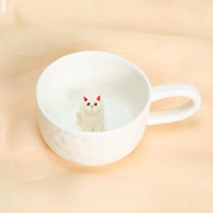 May include: White ceramic mug with a textured exterior and a sculpted white cat figurine inside. The cat has pink accents on its ears and nose. The mug has a curved handle and is set against a light background.