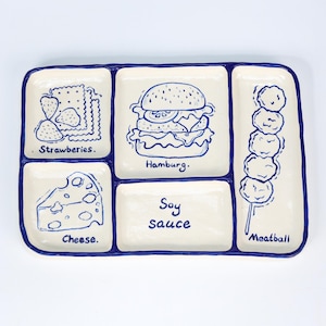 Könnte beinhalten: Ein rechteckiger, cremefarbener Keramikservierteller mit blauem Rand und fünf Fächern. Jedes Fach zeigt eine blaue Illustration und Text: Erdbeeren, Hamburger, Käse, Sojasauce und Fleischbällchen.