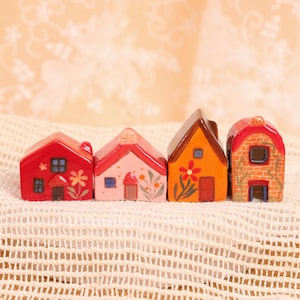 May include: Four miniature ceramic houses in a row, each with a unique design. The houses are painted in shades of red, pink, orange, and brick, with details like windows, doors, and floral accents. The houses are displayed on a white, textured surface.