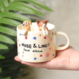K&ouml;nnte beinhalten: Cremefarbene Keramiktasse mit zwei Hundefiguren am Rand. Die Tasse ist mit bunten Blumenmustern und dem Text "NUBE & LINDT Amor eterno" verziert. Der Henkel hat die gleiche cremefarbene Farbe.