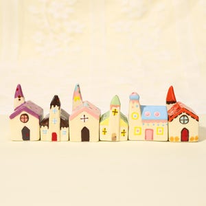 May include: A set of six miniature ceramic houses arranged in a row. Each house features a unique design with hand-painted details, including varied roof colors like purple, brown, pink, green, blue, and red, along with small doors and windows.