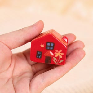 May include: A small, red ceramic house figurine with a whimsical design. The house features blue windows, a mushroom, and a flower. The figurine is held in a hand, showcasing its size and detail.