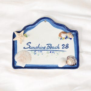 May include: A ceramic sign with a blue border and the words "Sunshine Beach 28". The sign is decorated with seashells and a starfish. The background is white.