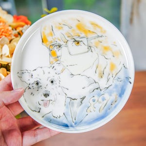Puede incluir: Un plato de cerámica blanco con un diseño pintado a mano de una persona con gafas y un perro. La persona tiene el pelo rubio y lleva una camisa azul. El perro es blanco y tiene la nariz negra. El plato tiene un borde azul y blanco.