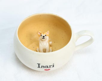 Puede incluir: Taza de cerámica color crema con una pequeña figura de Shiba Inu sentada en su interior. El interior es de color amarillo cálido. La palabra "Inari" está impresa en el exterior con un pequeño corazón rojo. La taza tiene un asa curvada.