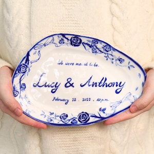 Op de afbeelding: Witte keramische serveerschaal met blauw bloemen- en lintontwerp. De schaal heeft de namen "Lucy & Anthony" en de datum "22 februari 2022, 22.00 uur" in blauw schrift. De tekst "We were meant to be" staat ook op de schaal.