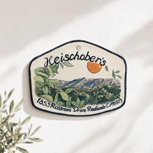 Op de afbeelding: Een crèmekleurige keramische plaquette met een donkerblauwe rand en een gat aan de bovenkant. Het toont een handgeschilderd landschap met groen gebladerte, blauwe bergen en een oranje zon. De tekst luidt Heischober's 1855 Rossmont Drive Redlands CA92373.