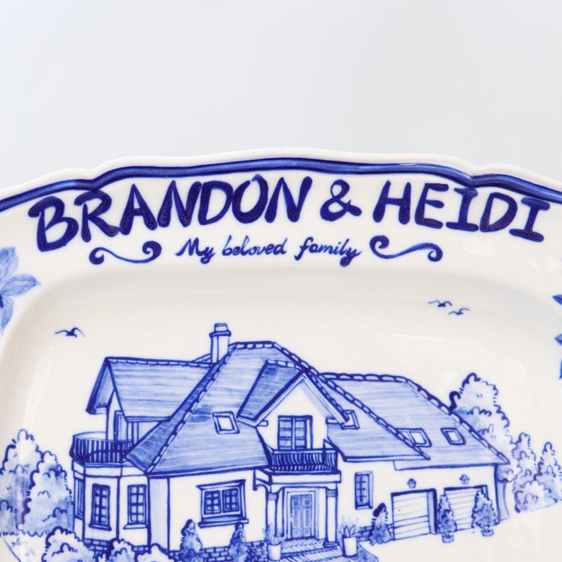 May include: A white ceramic serving platter with a blue hand-painted design. The platter features a house illustration and the text "BRANDON & HEIDI My beloved family". The rim has a scalloped edge with blue trim.