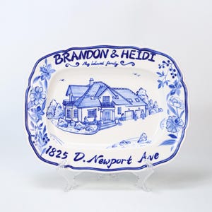 May include: A white ceramic serving platter with a blue floral border and a hand-painted illustration of a house. The platter is inscribed with the names "BRANDON & HEIDI", the phrase "My beloved family", and the address "1825 D. Newport Ave."