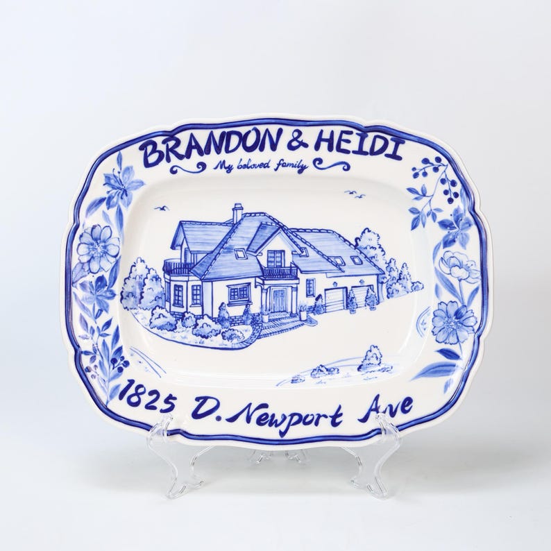 May include: A white ceramic serving platter with a blue floral border and a hand-painted illustration of a house. The platter is inscribed with the names "BRANDON & HEIDI", the phrase "My beloved family", and the address "1825 D. Newport Ave."