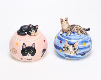 May include: Two decorative ceramic cat-themed jars. One is pink with a black cat face and a black cat figurine on top, labeled "Yogi." The other is blue with a tabby cat face and a tabby cat figurine, labeled "Marki."