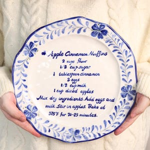 Puede incluir: Plato de cerámica blanca con diseño floral azul y la receta de Muffins de Manzana y Canela. La receta incluye ingredientes como harina, azúcar, canela, huevos, leche y manzanas en cubitos. Hornear a 190°C durante 20-25 minutos.